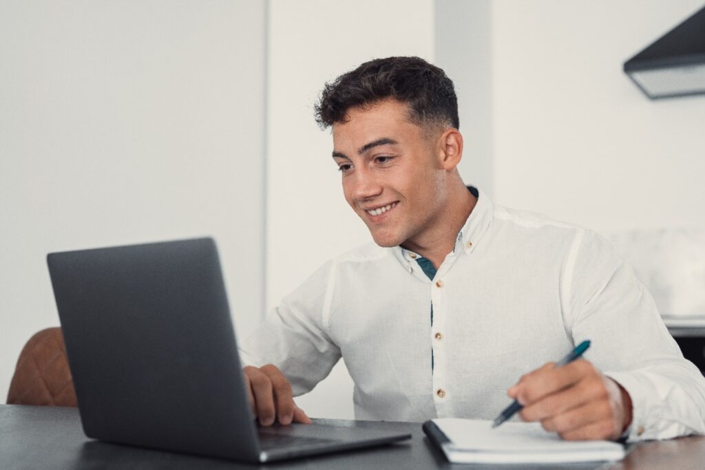 Jovem sorridente usando camisa social branca estuda com um notebook e anotações, representando preparação online para concursos públicos como o do DETRAN RJ.