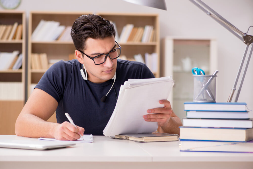 Homem jovem estudando com fones de ouvido e livros sobre a mesa, focado na leitura e anotações, representando preparação para concurso público.
