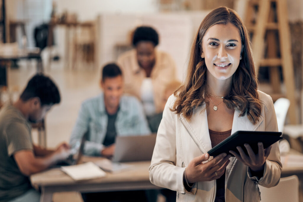 Mulher sorridente com tablet nas mãos em ambiente de trabalho colaborativo, representando uma profissional administrativa pronta para atuar no serviço público.